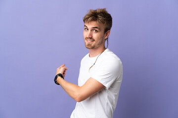 Young handsome blonde man isolated on purple background pointing back