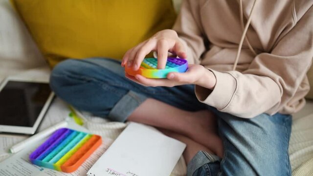 Teenage girl playing with rainbow pop-it fidget toy while studying at home. Teen kid with trendy stress and anxiety relief fidgeting game. Popping the dimples of sensory silicone toy.