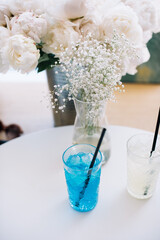Delicious refreshing blue and yellow lemonades in transparent plastic to-go cups standing on the white table with peony and gypsophila vase , vertical photo