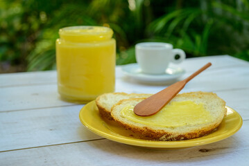 Ghee butter with sliced bread on yellow plate on breakfast table with glass jar of butter and cup...