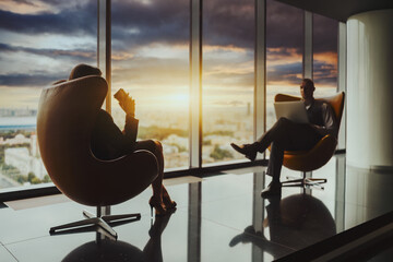Silhouettes of two businesspeople sitting in front of each other on bent chairs next to the window with a selective focus on a woman entrepreneur with a paper cup of coffee; a man with a laptop aloof - obrazy, fototapety, plakaty