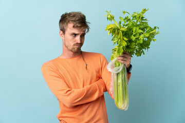 Young blonde man holding a celery isolated on blue background with sad expression