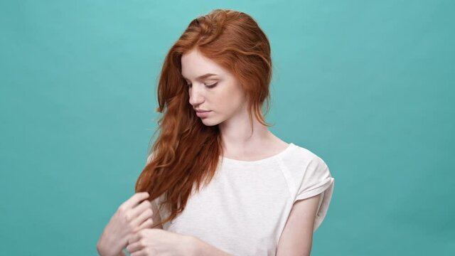 Smiling ginger woman in t-shirt touching her hair and looking around over turquoise background