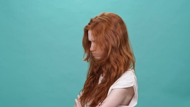 Offended serious ginger woman in t-shirt turned away with crossed arms over turquoise background