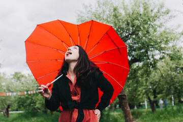 Pretty young woman carrying heart shaped umbrella and looking at camera with smile in the garden. Rain check