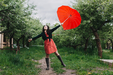 Pretty young woman carrying heart shaped umbrella and looking at camera with smile in the garden. Having fun under the rain