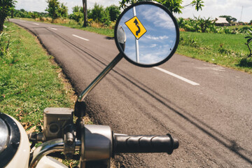 Scooter round mirror showing road curve traffic sign in countryside