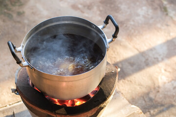 Cooking pork stew in a pot stainless steel is boiling and smoke on a charcoal stove close up.