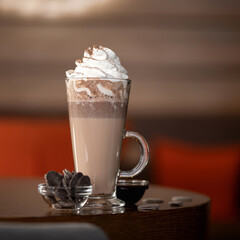 Long cup of macchiato coffee with whipped cream on top of it and glass bowl with chocolate cookies on blurred background with copy space for text. Square format.