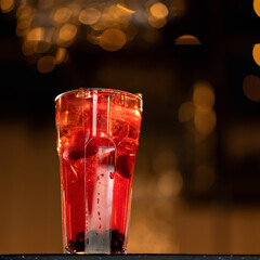 Glass of clear Red berry drink with ice cubes backlit on dark blurred background with bokeh....