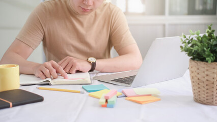 Student studying and writing notes in a notebook at home, making research on laptop and browse internet, watching online webinar, listening audio course, e-learning education concept