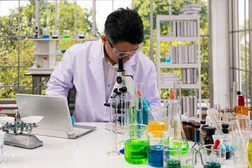 Senior scientist with safety glasses looking microscope and checking experimental data from laptop...