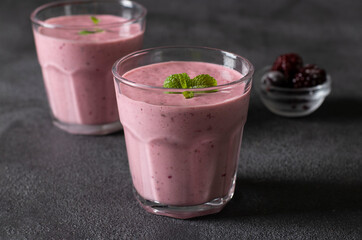 Homemade smoothies with frozen blackberries in two transparent glass on dark gray background.
