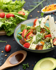 Bowl of colorful vegetable dish with microgreens on black table. Salad with red sturgeon fish and tomatoes. Close up shot. Soft focus. Vertical format.