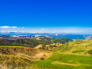Fototapeta na wymiar Green grass on the yellow spring alpine meadows of the Gumbashi pass. Snow covered huge mountain Elbrus on horizon against the blue sky. Mountainous, hilly summer landscape of the Caucasus Mountains.