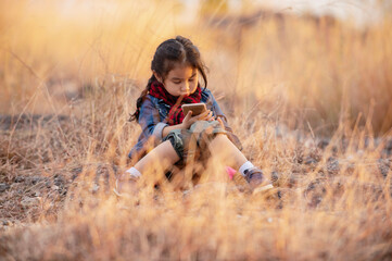 Naklejka na ściany i meble Adorable little girl uses mobile phone on meadow in summer sunset nature.