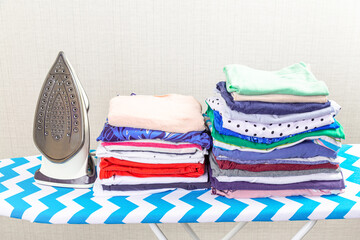 Electric iron on the ironing board and a pile of colorful clothes stacked on the ironing board