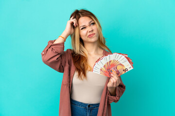 Teenager blonde girl taking a lot of Euros over isolated blue background having doubts and with...