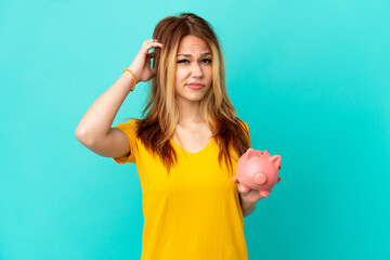 Teenager blonde girl holding a piggybank over isolated blue background having doubts