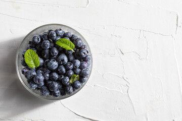 Delicious and appetizing blueberry berries in a glass bowl and mint leaves (peppermint, mint). On white textured background. Top view with copy space.