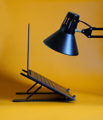 A modern slim silver laptop or ultrabook sits on a portable stand against a yellow background next to a desk lamp. Side view. Workplace concept