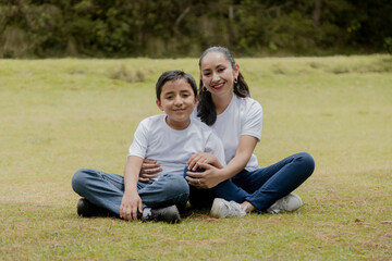 Fototapeta premium Young Hispanic mom with her son sitting in the park-mother hugging her son in the middle of the field outdoors