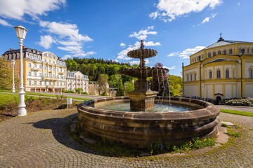 Naklejka premium Marianske Lazne, Czech Republic - May 30 2021: View of the baroque water fountain standing on Goethe square. Buildings, church and trees in the background. Sunny day in spa city. Blue sky and clouds. 