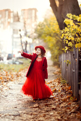 Cute funny caucasian child girl in a red coat walks on the autumn street, stylish child, a girl in a festive dress walking with yellow leaves, autumn mood, a child looks at the leaves of a tree