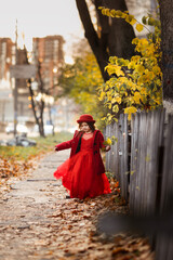 Fototapeta premium Cute funny caucasian child girl in a red coat walks on the autumn street, stylish child, a girl in a festive dress walking with yellow leaves, autumn mood, a child looks at the leaves of a tree