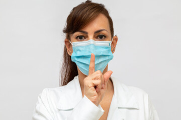 Enfermeira de uniforme e máscara cirúrgica azul, olhando para frente e com dedo, fazendo o gesto pedindo silêncio