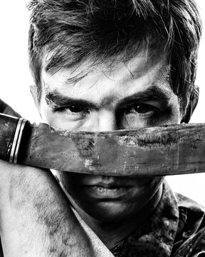 The soldier in bruises, shamray and combat coloring holds a machete in front of his eyes close-up. Black and white contrast portrait.
