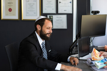 Obraz na płótnie Canvas Bearded jewish man in a Kippah sit office with lot of cash money. Bank loan or credit. Get cash in few minutes. Banking support line concept. Woman hands counts Israeli money, new shekels bills