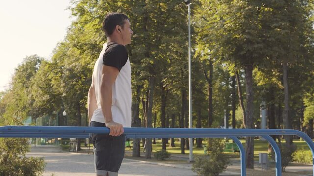 Young handsome guy doing physical exercise on the parallel bars. He listens to music on headphones.