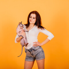 Gorgeous brunette woman smiling, holding Sphynx kitten, posing on grey and yellow studio background.