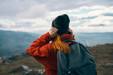 woman in a jacket hat and a backpack looks at the mountains in the distance in nature