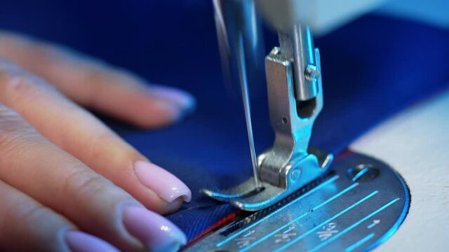 Female hand pushes material through a sewing machine. Sewing machine needle rapidly moves up and down. Tailor sews blue fabric in the sewing workshop. Close-up.