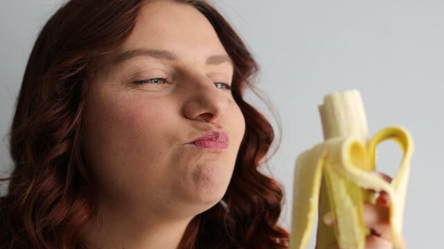Young woman laughing and eating ripe banana isolated on grey background. Woman biting banana, healthy eating and lifestyle