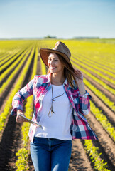 Smiling female farmer standing in soy field holding tablet and hat.