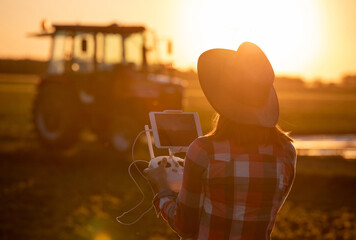 View from behind of female farmer using drone controller in front of tractor at sundown. © Budimir Jevtic