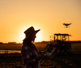 Agronomist using drone in agriculture standing in field in front of tractor at sundown.