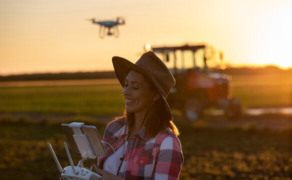 Female Farmer Flying Drone Over Field Using Controller With Tractor In Background.