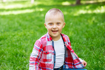 Happy boy with Down syndrome in the park against a background of green grass, close-up portrait of...