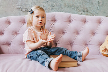 little girl 1 year old blonde with blue eyes sitting in jeans on a pink sofa with a book, an early...