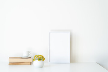 Empty white picture frame mockup in sunlight. Books, coffee cup and sphere-shaped vase with small yellow flowers. Concept of waking up and good morning with optimism White table and wall in background