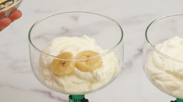 Whipped cream cheese desert with banana slices in dessert glasses close up on marble kitchen table, woman hands
