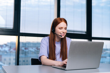 Close-up of friendly young woman operator using headset and laptop during customer support at home office. Young female student communicating online by video call on background of window.