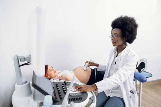 Young likable professional attractive African American woman doctor doing 3d ultrasound on belly of pregnant woman in clinic. Ultrasound for pregnant woman.