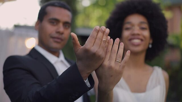 Close-up male and female African American hands with wedding ring on finger and blurred happy excited couple of newlyweds at background. Cheerful bride and groom bragging jewelry on marriage ceremony