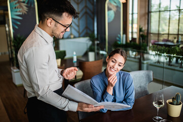 Hansome young waiter showing menu to beautiful female customer in restaurant.