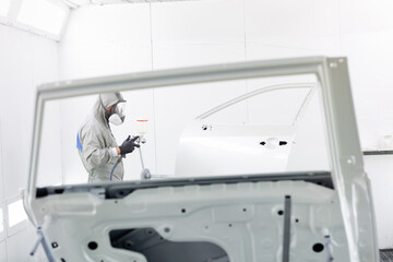 Obraz premium Man in overalls paints the car door using factory technology. garage for car painting. man in a gray spacesuit. doors for the car on repair of white color